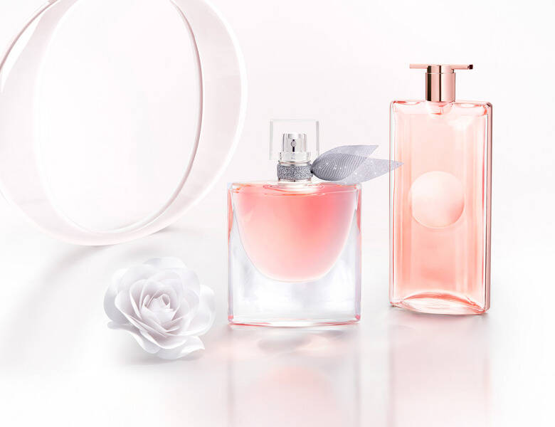 Her ses et billede med to parfumeflakoner i lyserøde nuancer placeret på en lys, reflekterende overflade mod en lys baggrund. Den ene flakon har en sløjfedetalje ved halsen. Ved siden af flakonerne ses en hvid blomst samt dekorative, runde elementer i baggrunden.