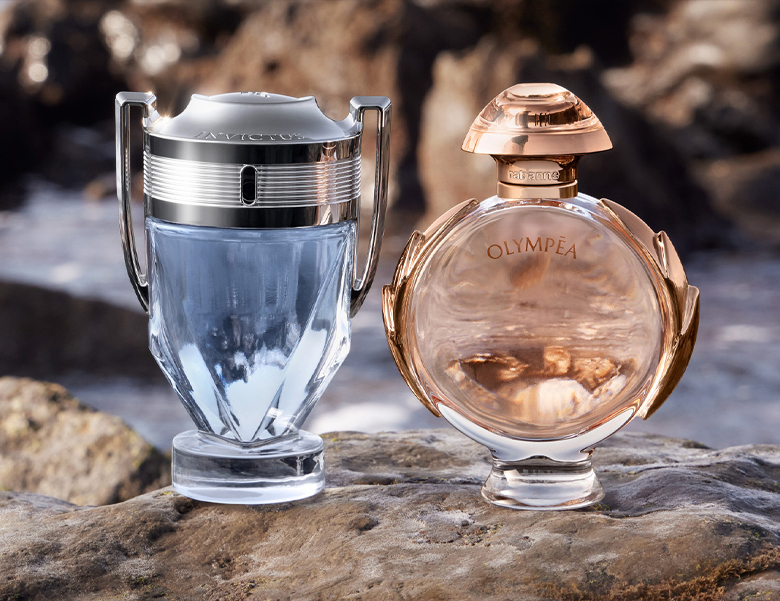 Her ses et billede med to parfumeflakoner placeret side om side på sten ved vand. På den ene flakon står “INVICTUS”, og på den anden står “OLYMPÉA”. Flakonerne har henholdsvis sølv- og guldfarvede detaljer.