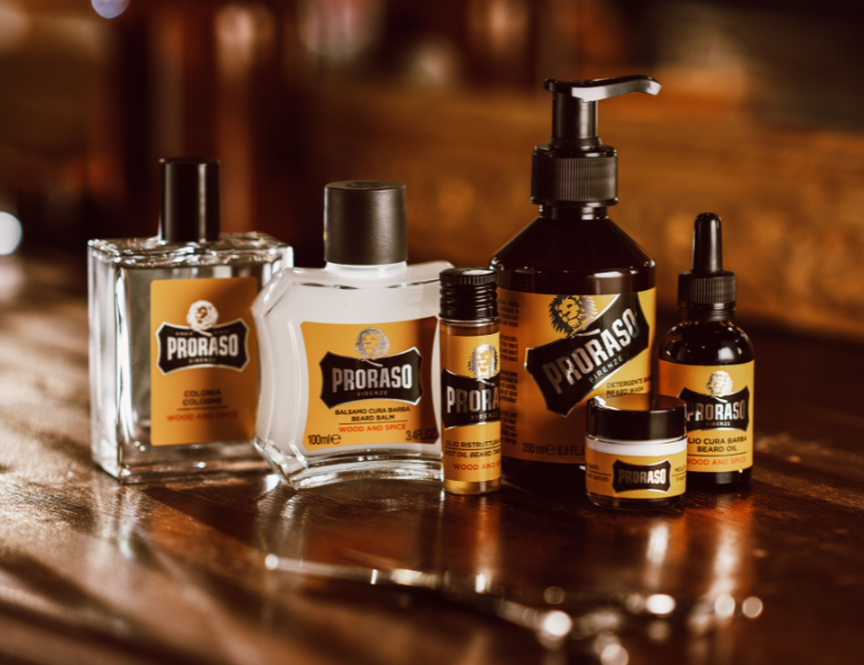 Proraso skægplejeprodukter står samlet på et mørkt træbord i varm belysning, herunder cologne, balsam, shampoo, olie og voks.