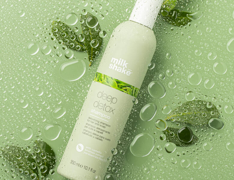 Her ses et billede af en shampoo-flaske fra milk_shake placeret mod en grøn baggrund med vanddråber og blade. På flasken står teksten “milk_shake deep detox shampoo”. Flasken er i lysegrøn og hvid emballage.