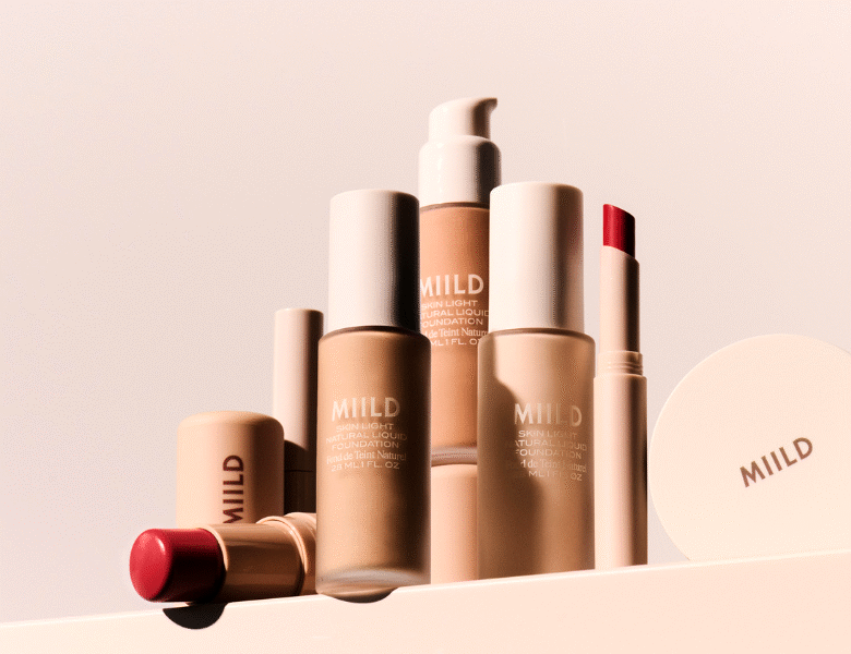 Her ses en samling af makeup-produkter fra MIILD placeret på en lys, neutral baggrund. Produkterne inkluderer flasker med foundation, en pumpeflaske, en stick og en rund kompakt, alle i beige og neutrale nuancer med “MIILD” skrevet på emballagen.