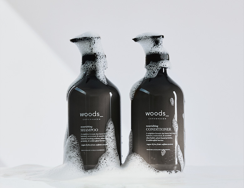 Her ses WOODS_ shampoo og conditioner i mørke flasker med skum på en lys baggrund.