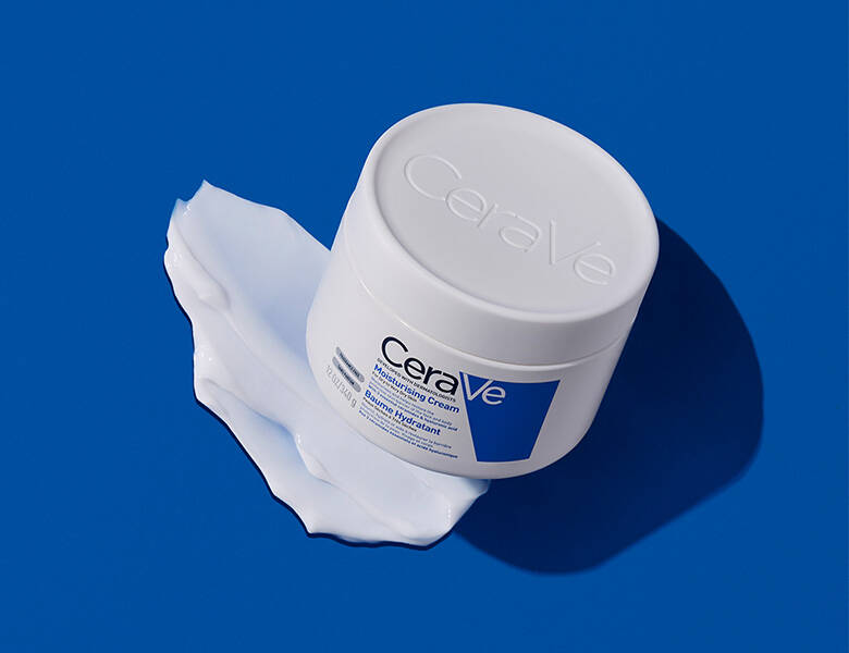 En rund, hvid krukke fra CeraVe står på en blå baggrund. Låget har præget logo, og krukken står oven på en udtværet mængde hvid creme, som breder sig ud under den. Billedet har fokus på produktets tekstur og en ren, klinisk æstetik.