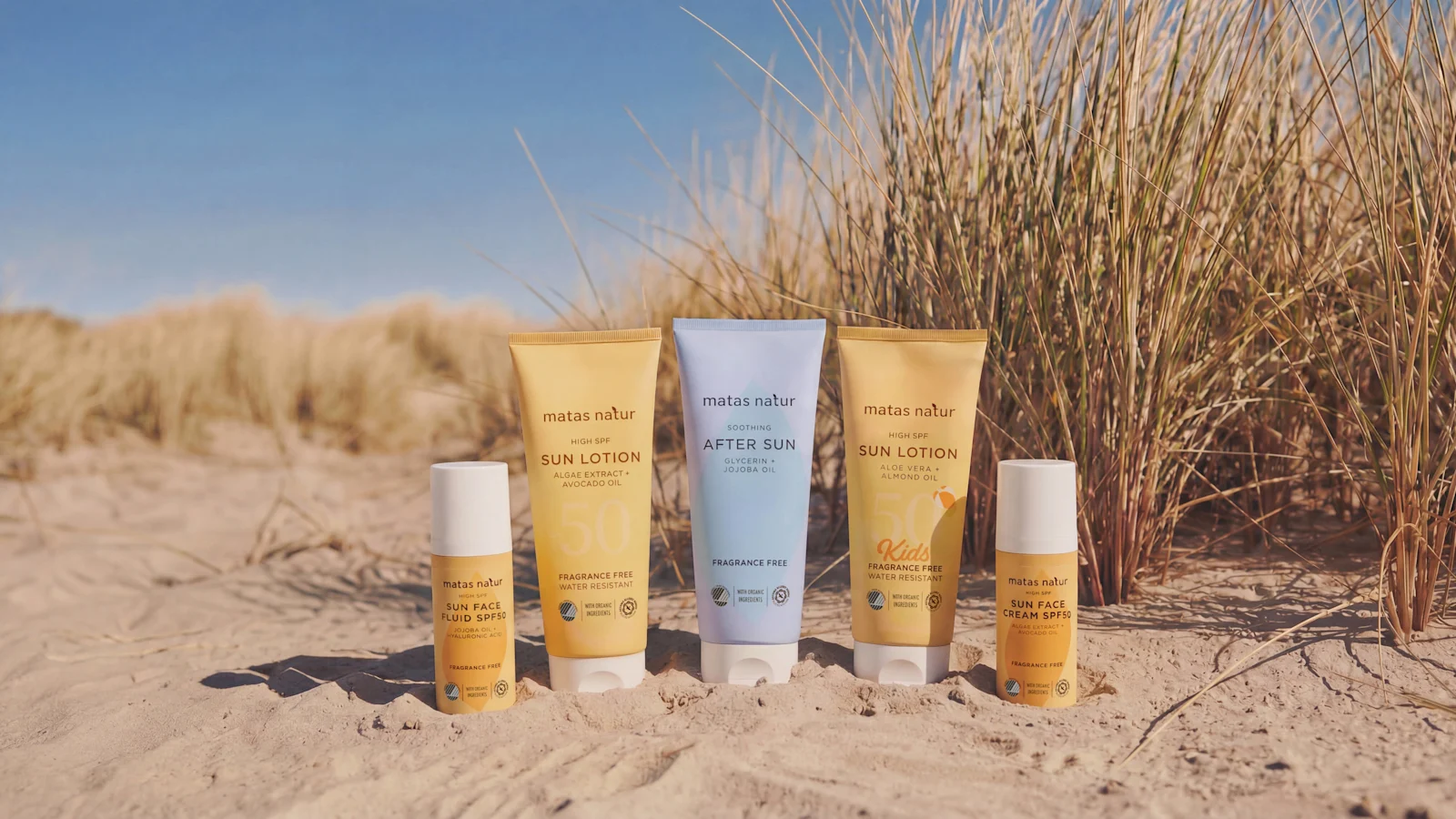 Her ses Matas Natur solpleje med Sun Face Fluid SPF 50, Sun Lotion SPF 50, Kids Sun Lotion SPF 50, After Sun og Sun Face Cream SPF 50 placeret i sandet foran marehalm på en strand.