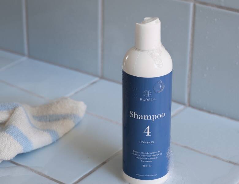 Her ses et billede af en shampoo-flaske placeret på en lys overflade ved en væg med lyse fliser. På flasken står teksten “PURELY Shampoo 4”. Der ses en klud ved siden af flasken, og der er vanddråber på produktet.
