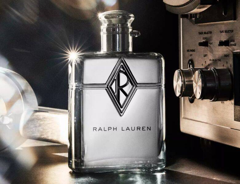 Her ses et billede af en parfumeflakon med teksten “RALPH LAUREN” på fronten. Flakonen er gennemsigtig med et markeret logo på midten og placeret på en mørk overflade. I baggrunden ses teknisk udstyr med knapper og drejeknapper samt lysreflekser.