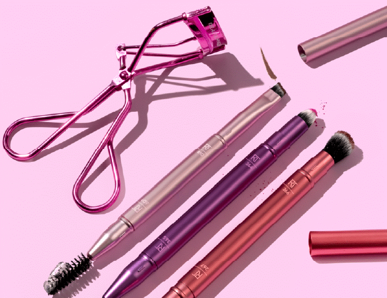 Her ses en lyserød baggrund, hvorpå der ligger fem Real Techniques makeupbørster og en øjenvippebukker. Alle produkterne har lyserøde og lilla farver. 