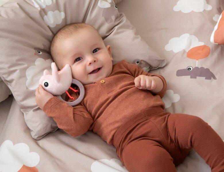 Her ses en glad baby med et legetøj i hånden. Babyen ligger i en blød seng. 