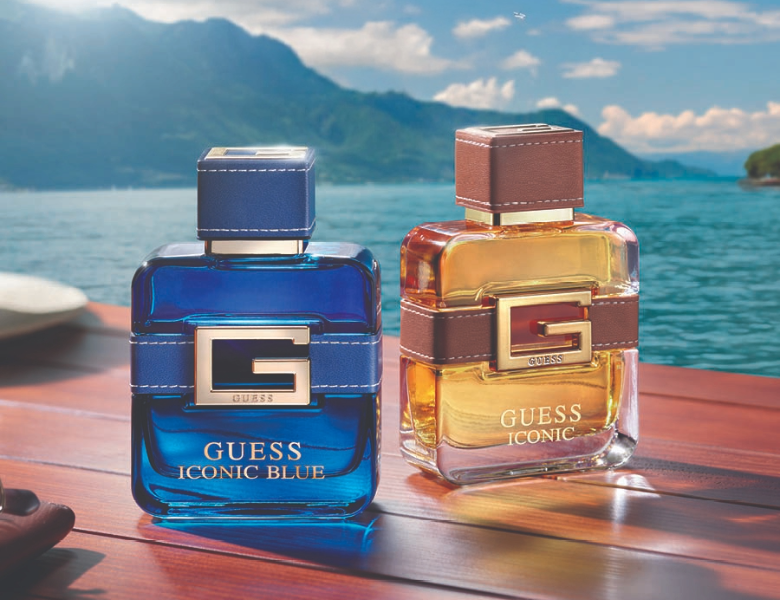 Her ses et billede med to parfumeflakoner fra Guess placeret på et træbord ved vandet med bjerge i baggrunden. På flakonerne står teksten “GUESS ICONIC BLUE” og “GUESS ICONIC”. Flakonerne er i blå og gyldne nuancer med store “G”-detaljer på fronten.