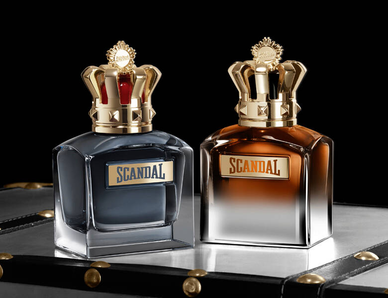 Her ses et billede med to parfumeflakoner med teksten “SCANDAL” på fronten. Den ene flakon er mørkeblå, og den anden er brunlig med en gradient mod klar bund. Begge har guldfarvede låg formet som kroner. Produkterne er placeret på en mørk overflade med dekorative kuffertelementer mod en sort baggrund.