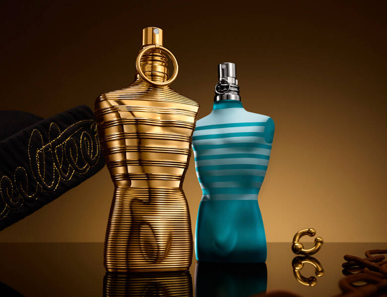 Her ses et billede med to parfumeflakoner formet som overkroppe placeret side om side. Den ene flakon er guldfarvet med vandrette linjer, og den anden er turkis med lyse striber. Produkterne står på en reflekterende overflade mod en gylden baggrund, og der ses dekorative elementer omkring dem.