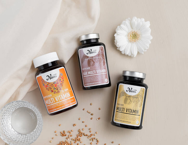 Tre mørke Nani bøtter med multivitaminer står på lys baggrund sammen med en hvid blomst, et glas vand, lyst stof og små spredte frø.