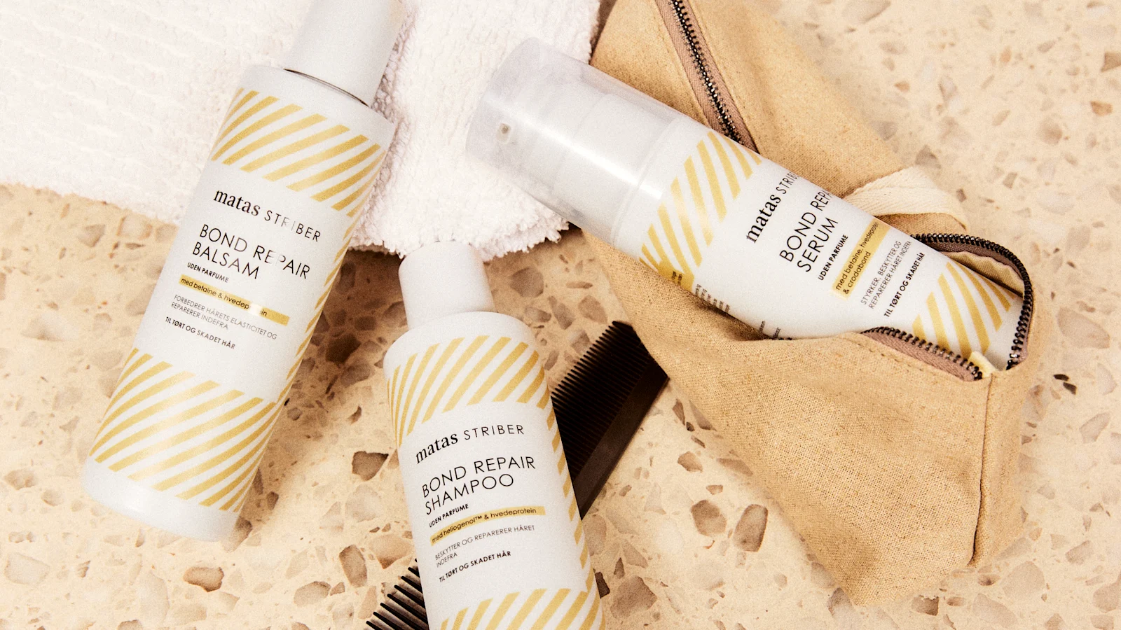 Her ses Matas Striber Bond Repair Shampoo, Bond Repair Balsam og Bond Repair Serum på et terrazzobord sammen med et håndklæde, en toilettaske og en kam.