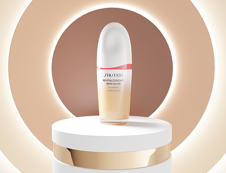 Her ses et billede af et foundation-produkt fra Shiseido placeret på et podium mod en beige baggrund med cirkulært lys. På produktet står teksten “SHISEIDO REVITALESSENCE SKIN GLOW Foundation”.