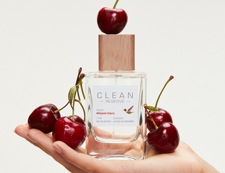 Parfumeflaske fra CLEAN RESERVE “Whipped Cherry” holdt i en hånd omgivet af røde kirsebær.