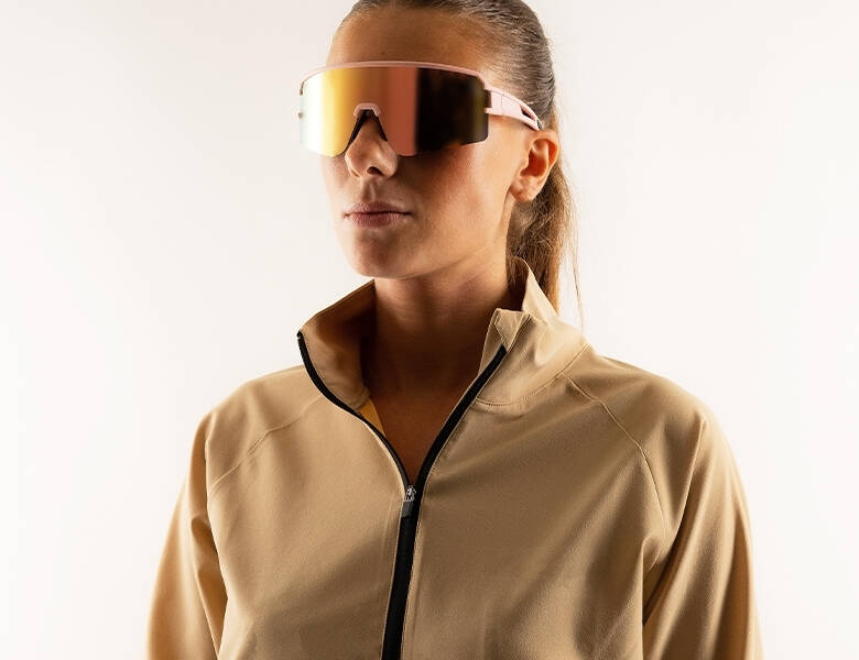 Her ses en hvid baggrund med en kvinde i beige tøj. Hun har et par sportssolbriller på.