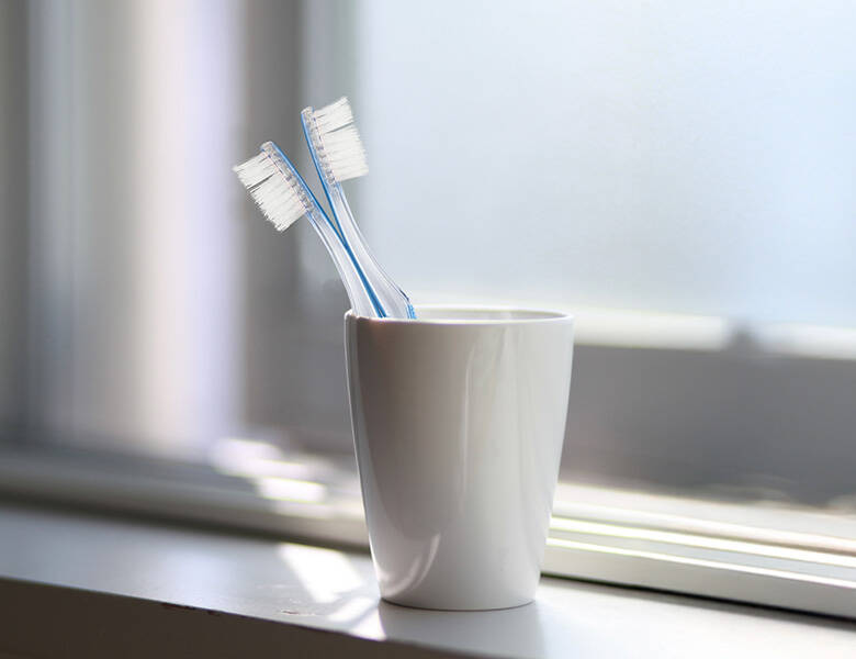 Her ses et hvidt krus, som står i en hvid vindueskarm. Kruset har to tandbørster i sig. 