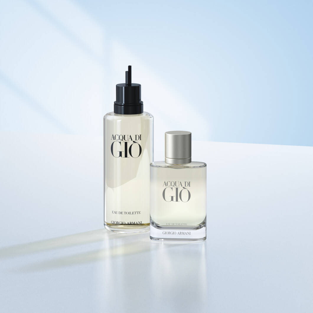 To Acqua di Giò-parfumeprodukter fra Giorgio Armani står på en lys overflade foran en blød lyseblå baggrund. Til venstre ses en høj refillflaske med sort pumpe, og til højre står den klassiske parfumeflaske med sølvfarvet låg. Billedet har et rent og elegant udtryk med fokus på luksusparfume og refill-løsninger.