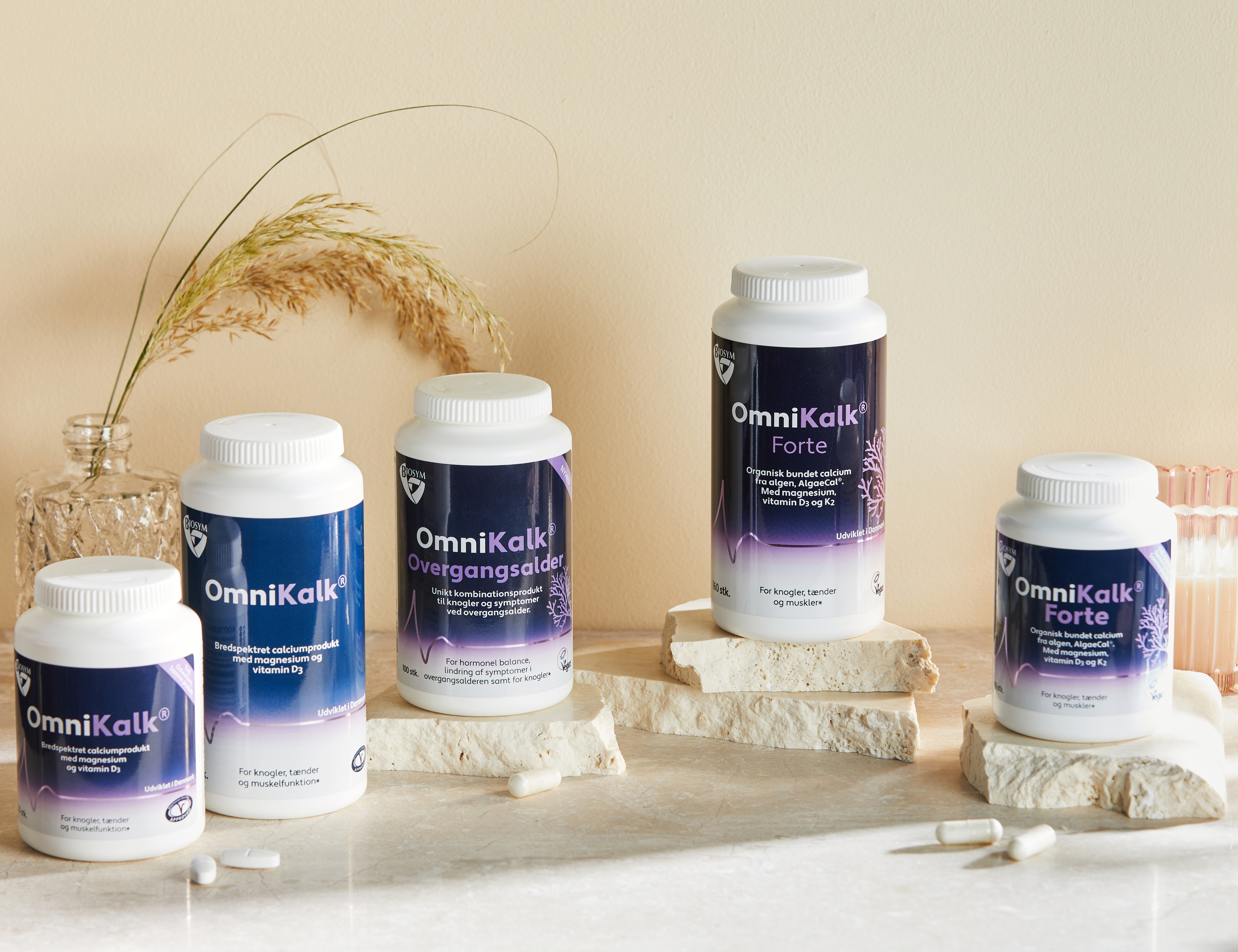 Flere OmniKalk kosttilskud står på lyse stenplader på en beige baggrund sammen med kapsler, en glasvase med tørrede strå og et lyserødt glas.