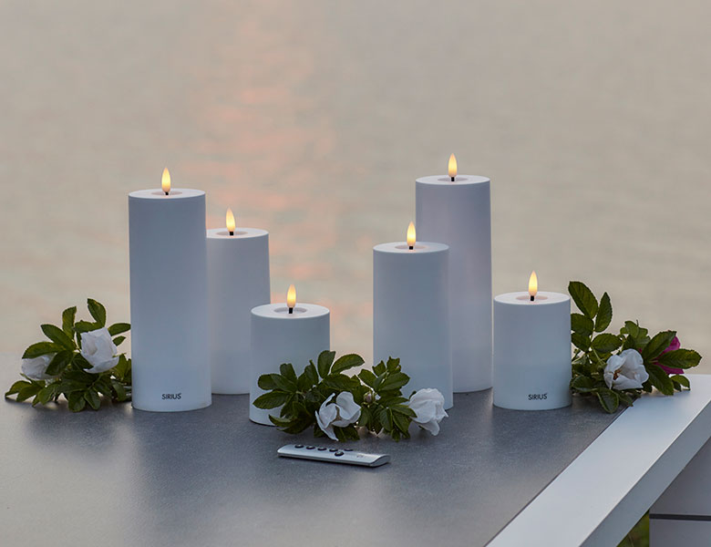 Her ses hvide LED-lys med flammeeffekt omgivet af blomster på et bord udendørs.