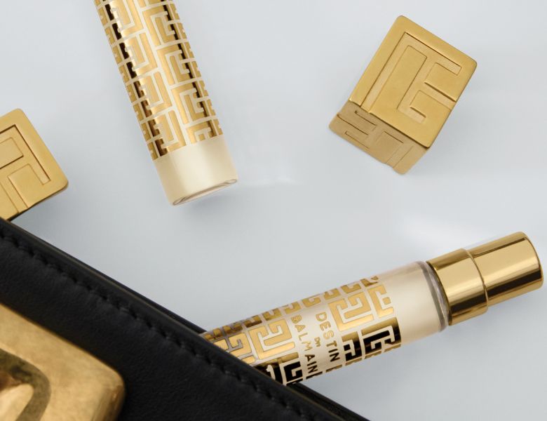 Her ses et billede med mindre parfumeflakoner fra Balmain placeret mod en lys baggrund. Produkterne har guldfarvede og lyse nuancer med mønstre, og der ses også guldfarvede låg og detaljer omkring dem.
