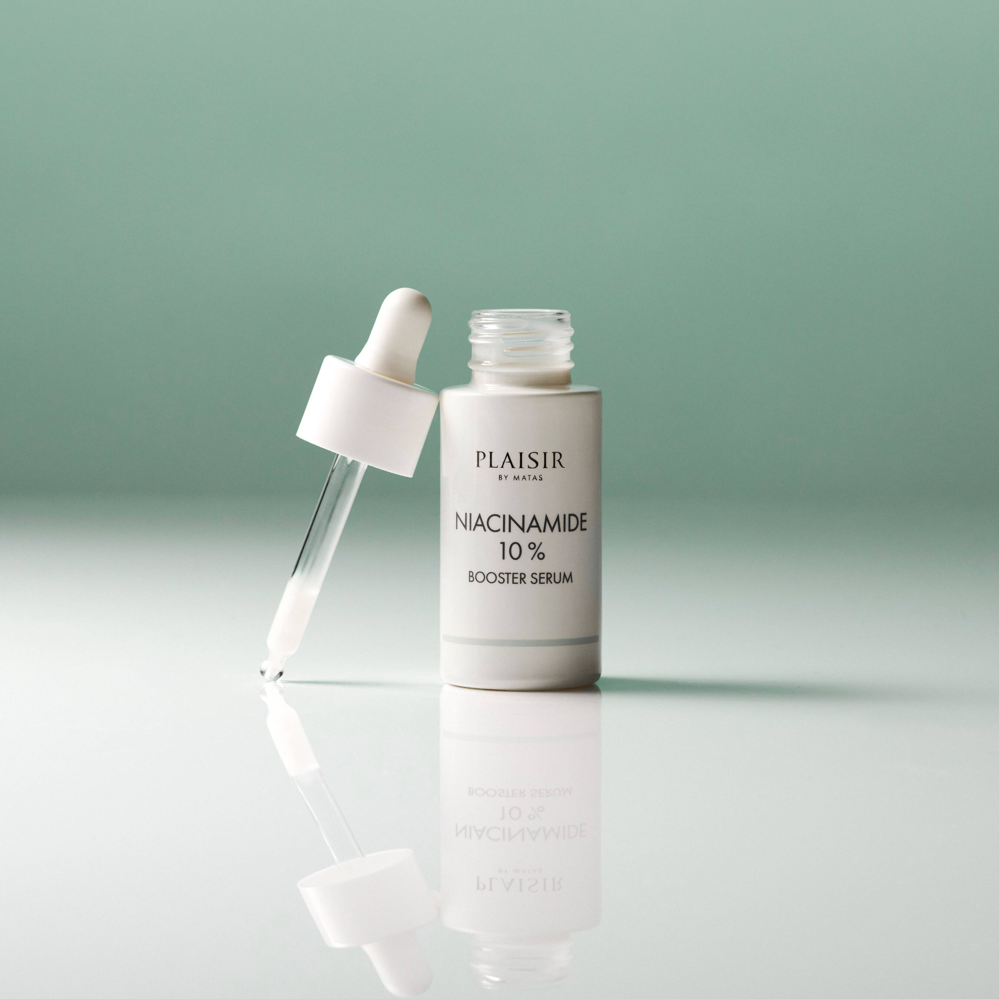 En flaske Plaisir Niacinamide 10% Booster Serum står åben på en blank overflade mod en grøn baggrund. Pipetten ligger ved siden af med en dråbe i spidsen, og flasken spejler sig i underlaget.