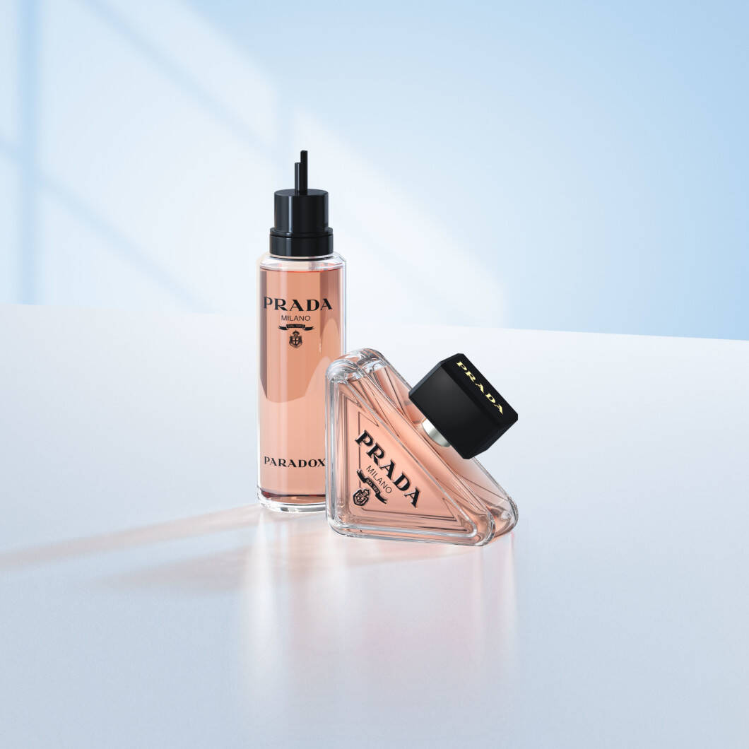 To Prada Paradoxe-parfumeprodukter står på en lys overflade foran en blød lyseblå baggrund. Til venstre ses en høj refillflaske med sort pumpe og lyserød parfume. Til højre ligger den ikoniske trekantede Prada Paradoxe-parfumeflaske med sort låg. Billedet har et elegant og moderne udtryk med fokus på luksusparfume og refill-løsninger.