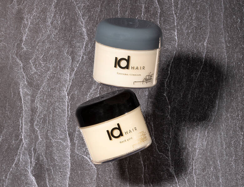 Her ses to forskellige IdHAIR hårvoks liggende på et mørkt marmorbord. Den ene voks har et sort låg og den anden et gråt.