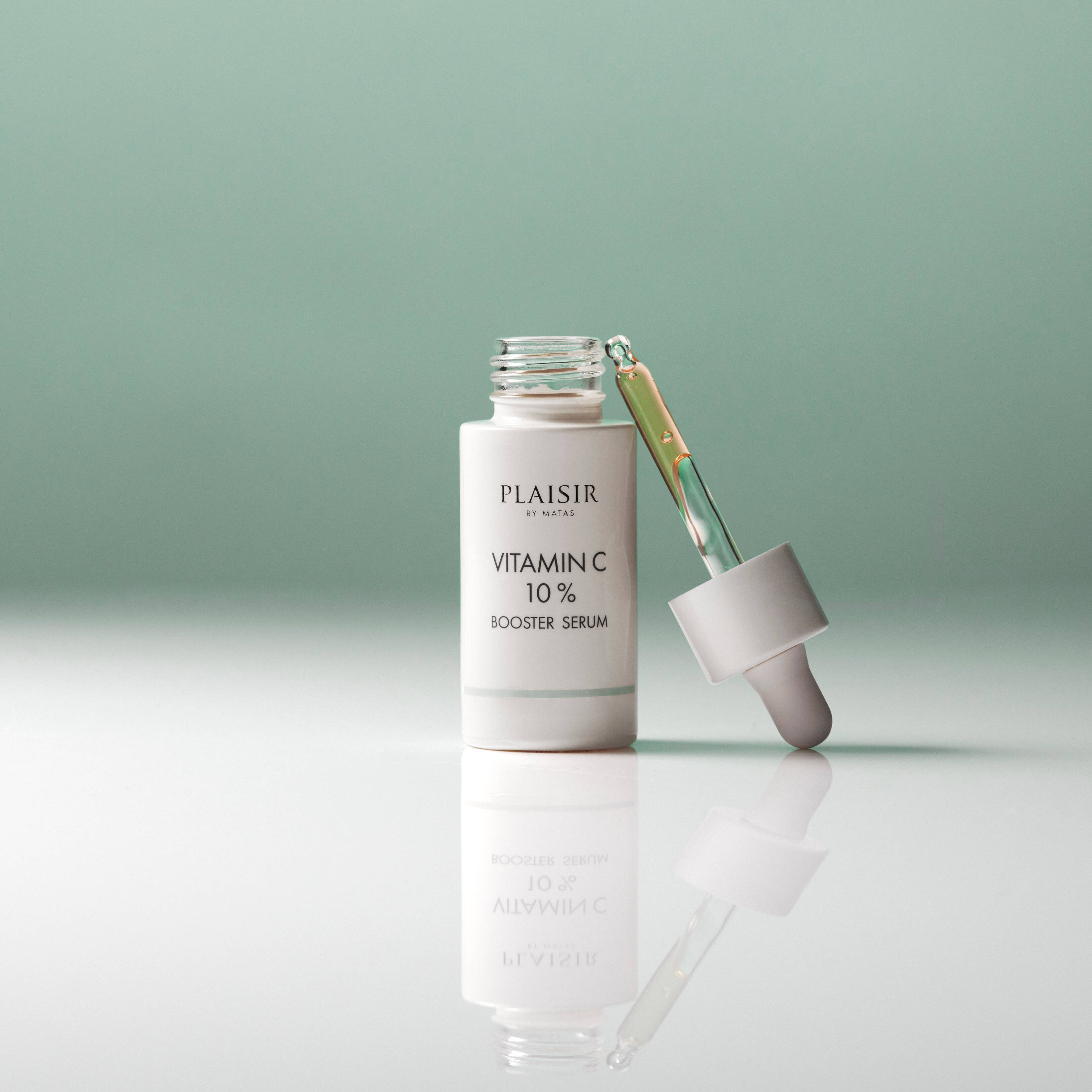 En flaske Plaisir Vitamin C 10% Booster Serum står åben på en blank overflade mod en grøn baggrund. Pipetten med gylden serum ligger ved siden af, og flasken spejler sig i underlaget.