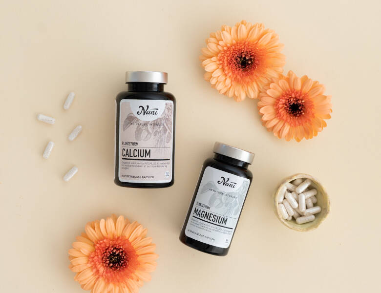 To mørke Nani bøtter med calcium og magnesium ligger på beige baggrund omgivet af orange blomster, hvide kapsler og en lille skål med kapsler.