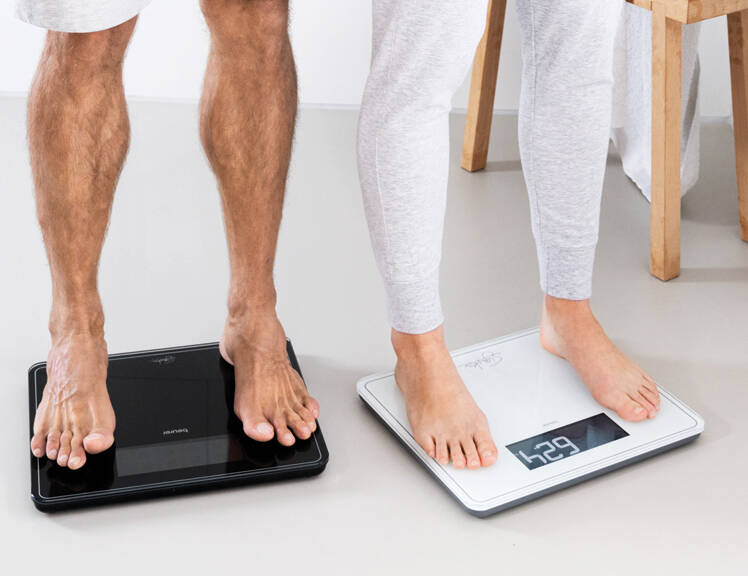 Her ses benene af to mennesker, som hver står på en badevægt - en i hvid og en i sort. 