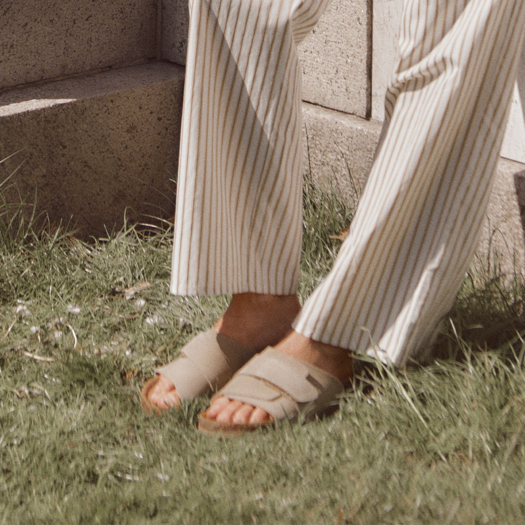 Her ses et par fødder iført sandaler og stribede bukser på græs ved en mur.
