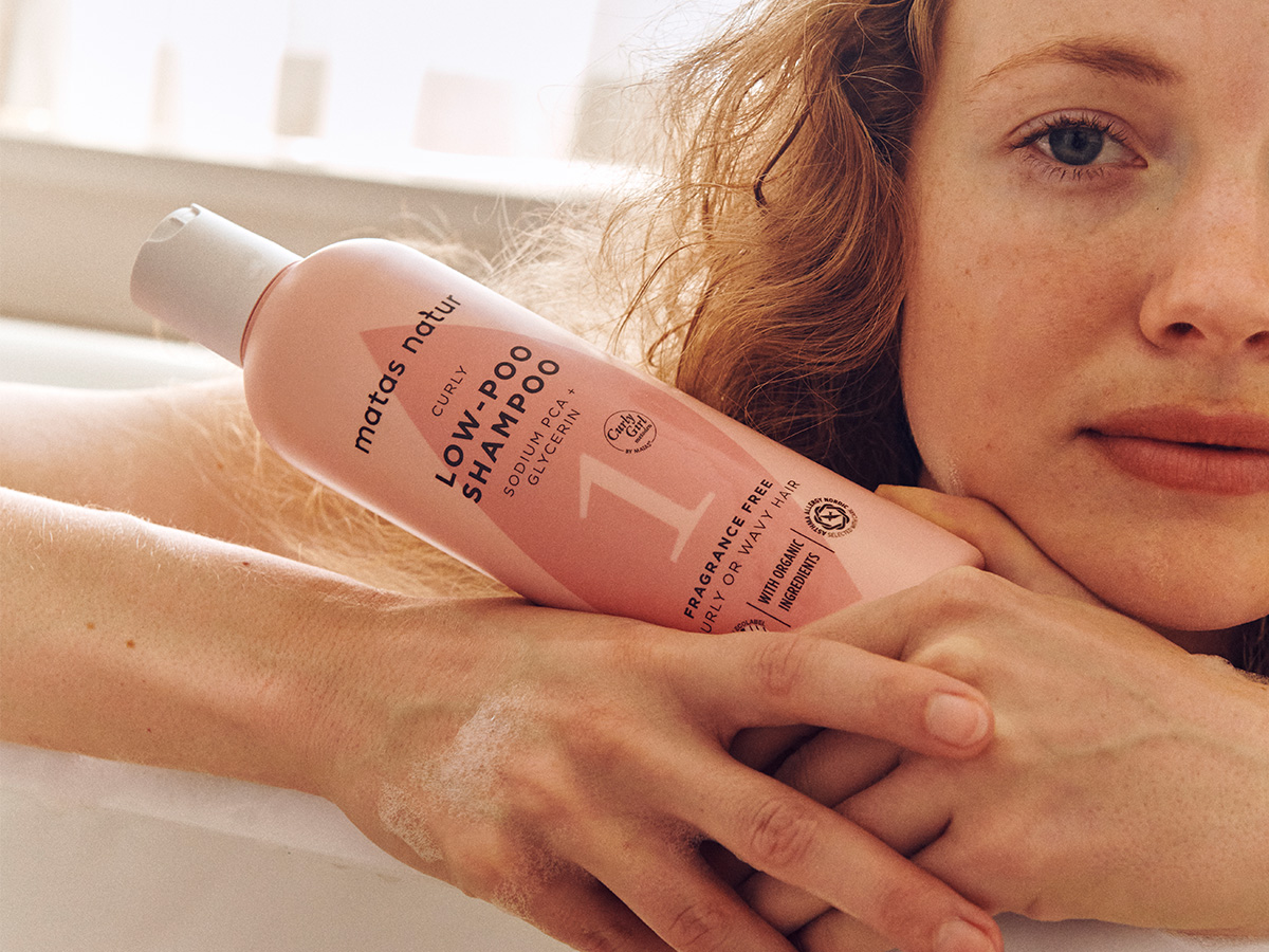 Her ses en person med krøllet rødt hår, som holder en rosa flaske Matas Natur Curly Low-Poo Shampoo tæt ved ansigtet. På flasken står blandt andet “Sodium PCA + Glycerin”, “Fragrance Free” og “Curly or Wavy Hair”.