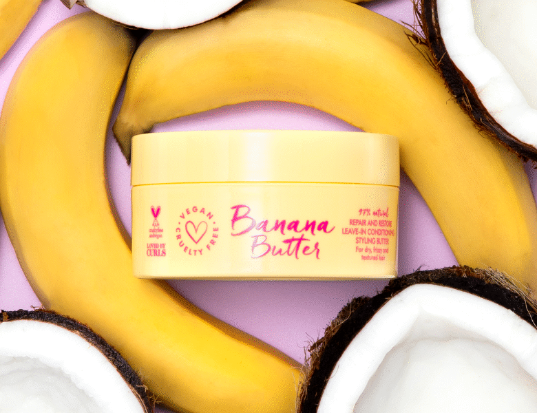 Her ses et billede af et hårprodukt med teksten “Banana Butter” placeret centralt mod en lyserød baggrund. Produktet er omgivet af bananer og kokosstykker. På emballagen står også “VEGAN" og “LEAVE-IN CONDITIONER”.