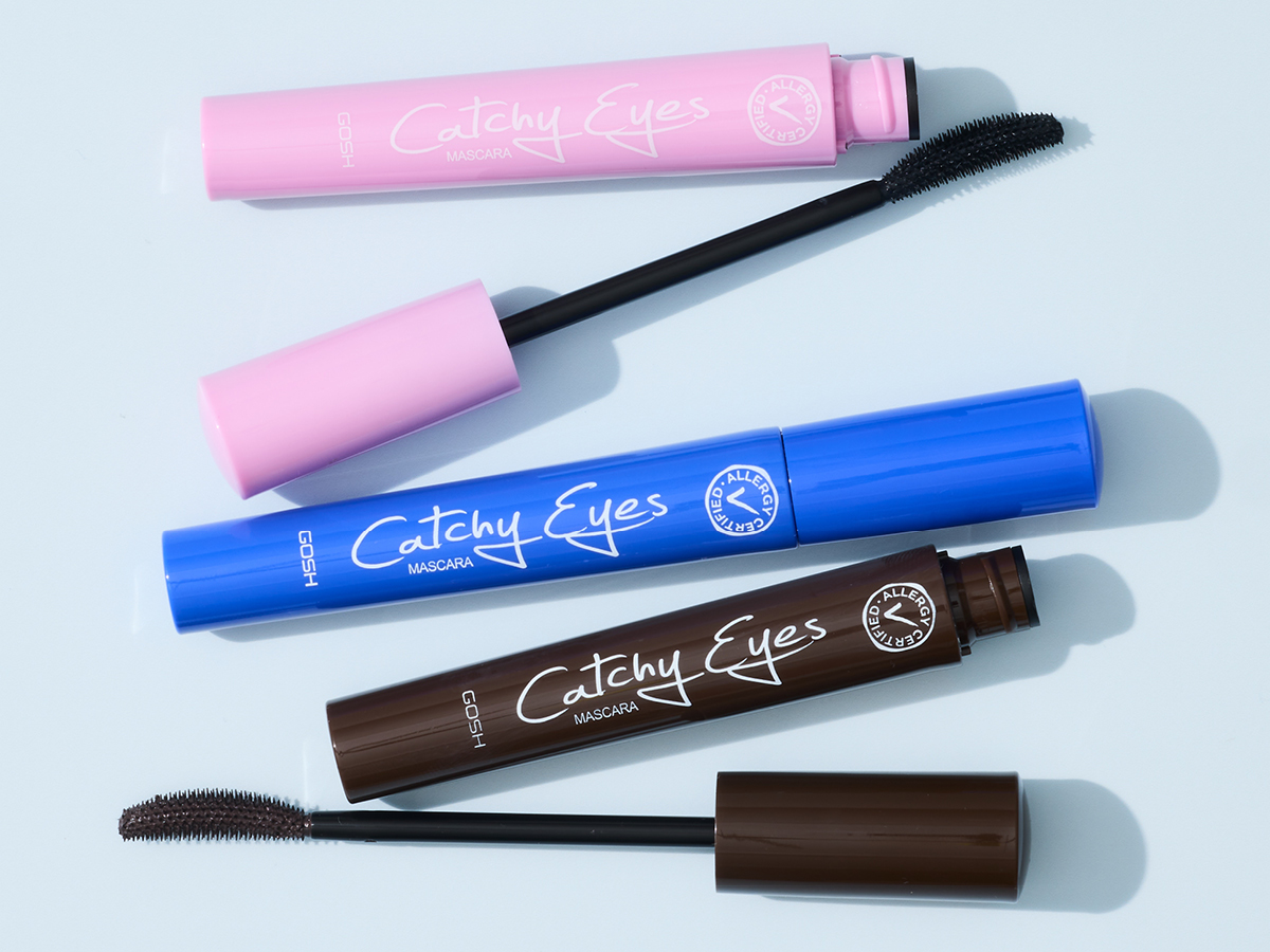 Her ses flere Catchy Eyes Mascara fra GOSH Copenhagen i pink, blå og brun emballage mod en lyseblå baggrund. To mascaraer er åbne, så de sorte mascarabørster er synlige.