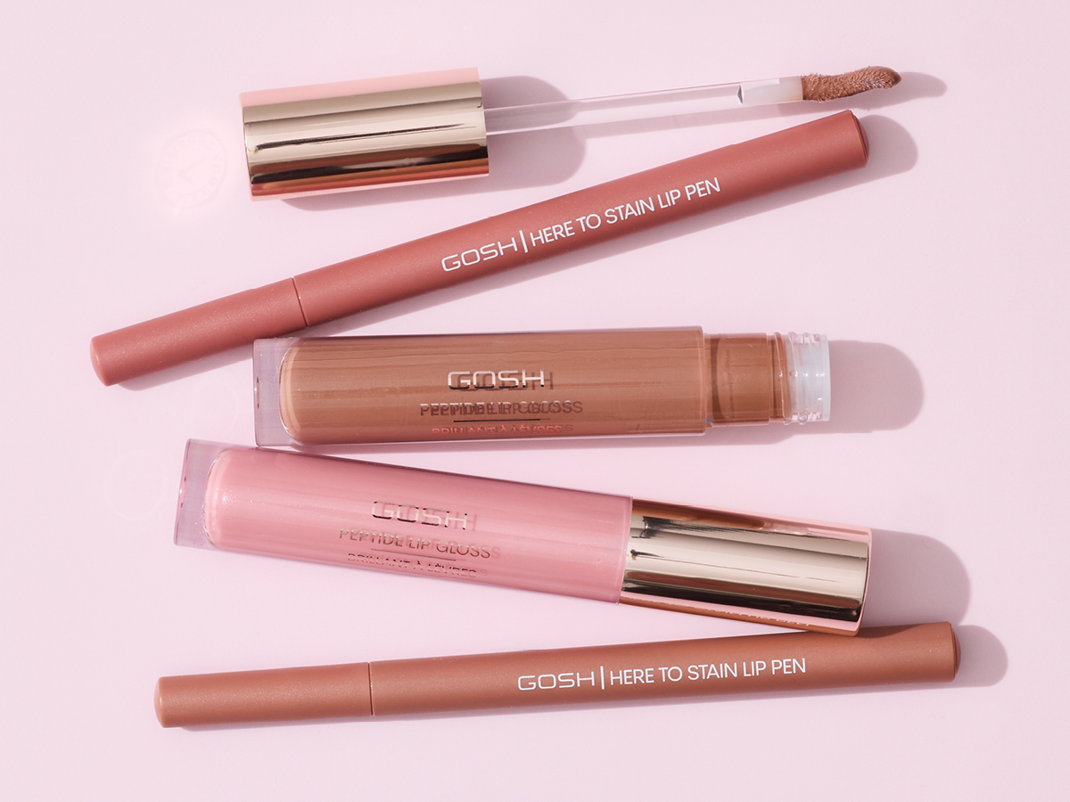Her ses et udvalg af læbeprodukter fra GOSH Copenhagen mod en lyserød baggrund. Produkterne omfatter Here to Stain Lip Pen og Peptide Lip Gloss i forskellige rosa og nude nuancer.