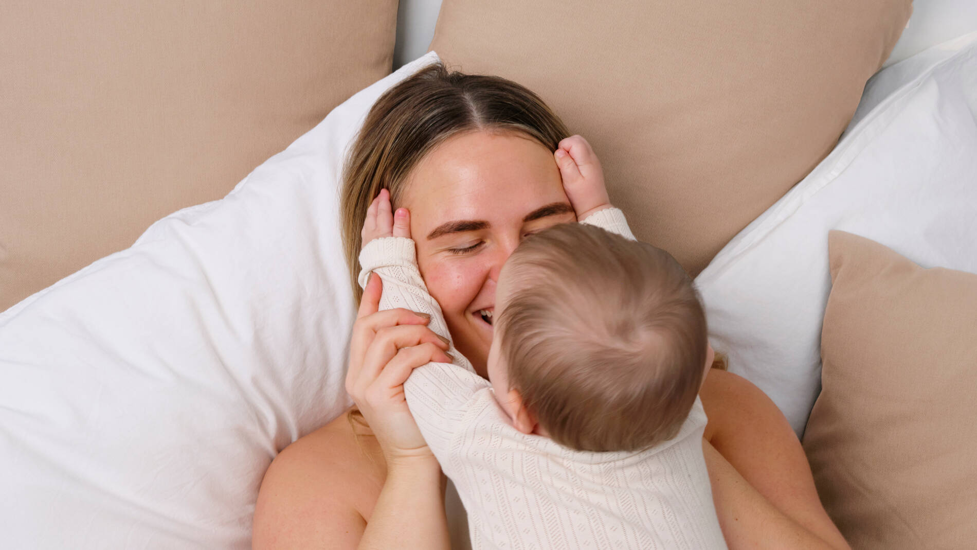 En voksen og et lille barn ligger tæt sammen i en seng omgivet af store hvide og beige puder. Barnet holder sine hænder på den voksnes ansigt og læner sig ind til et kærligt kram, mens den voksne smiler med lukkede øjne. Begge har lyst tøj på, og billedet har en varm, rolig og nærværende stemning.
