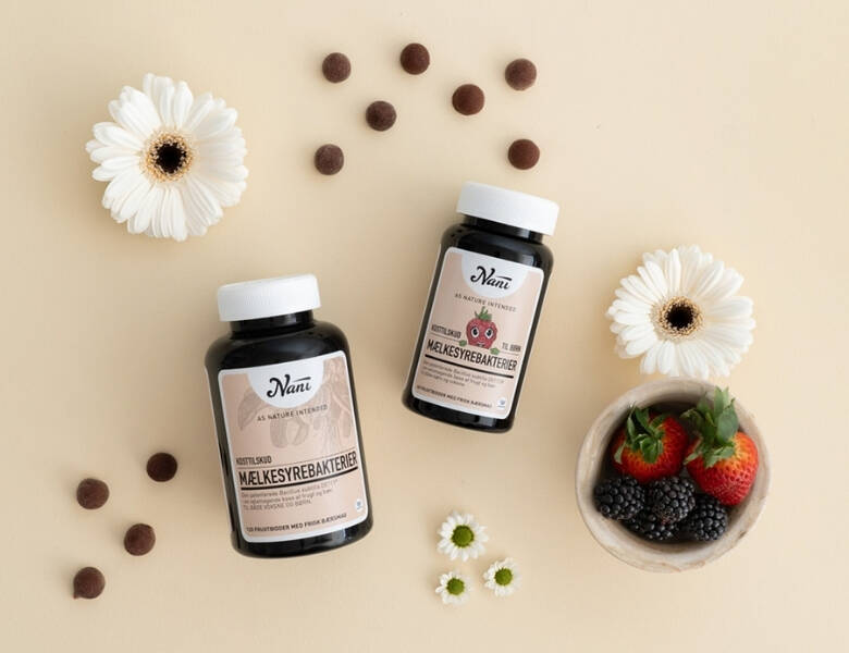 To mørke Nani bøtter med mælkesyrebakterier ligger på beige baggrund omgivet af hvide blomster, små brune tabletter og en skål med jordbær og brombær.
