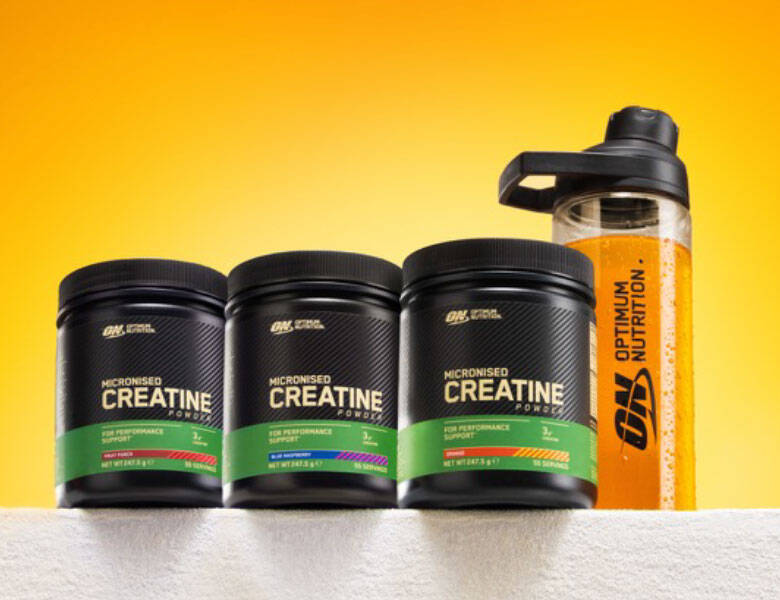 Billedet viser tre sorte bøtter med Optimum Nutrition Micronised Creatine Powder (flavoured creatine monohydrat) med grønne etiketter og forskellige smagsindikatorer, placeret side om side på en lys overflade. Til højre står en klar træningsshaker med Optimum Nutrition-logo fyldt med orange væske. Baggrunden er en jævn gul-orange gradient, der fremhæver produkterne.