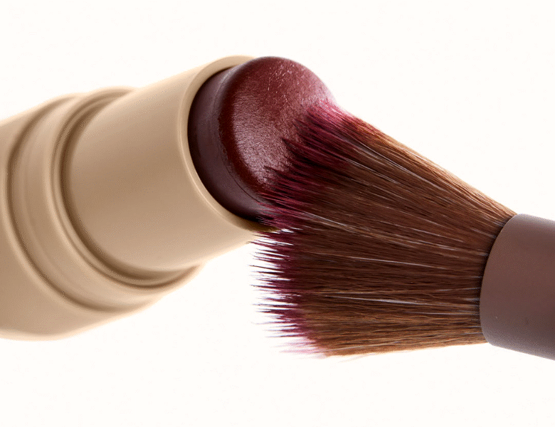 Her ses et nærbillede af en blush-stick i en beige emballage, hvor en makeupbørste påfører produktet fra overfladen. Produktet har en dyb rød nuance, og baggrunden er lys og neutral.