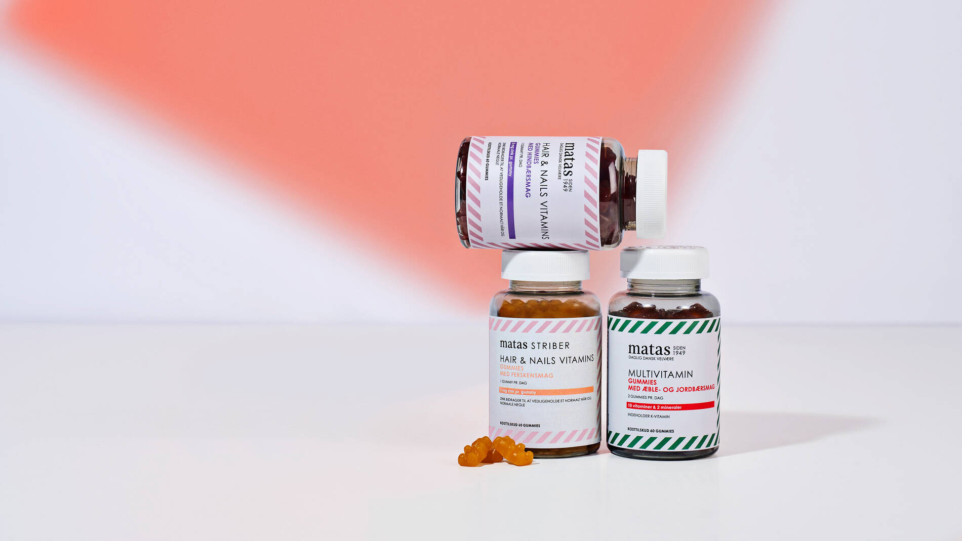 Tre glas med Matas vitaminer og gummies står opstillet på en hvid overflade foran en baggrund med en blød koralfarvet skygge. To glas står oprejst side om side, mens et tredje glas ligger vandret ovenpå dem. Et af de stående glas indeholder multivitamin-gummies med grøn- og hvidstribet etiket, og det andet indeholder hair & nails-vitaminer med lyserødstribet etiket. Foran glassene ligger et par orange gummies formet som små bjørne.