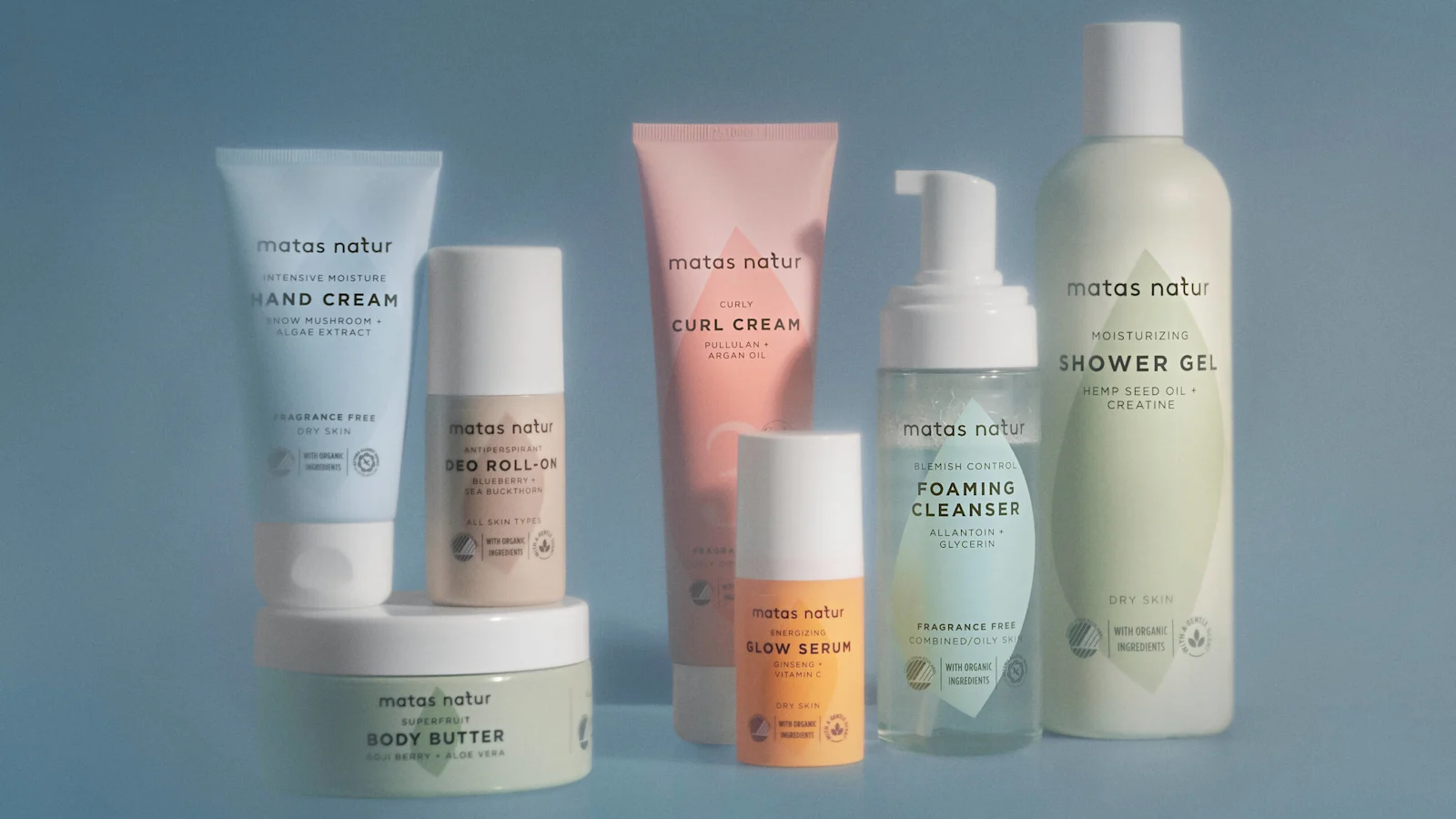 Her ses et udvalg af Matas Natur produkter, herunder Hand Cream, Body Butter, Deo Roll-On, Curl Cream, Glow Serum, Foaming Cleanser og Shower Gel, opstillet på en lyseblå baggrund.