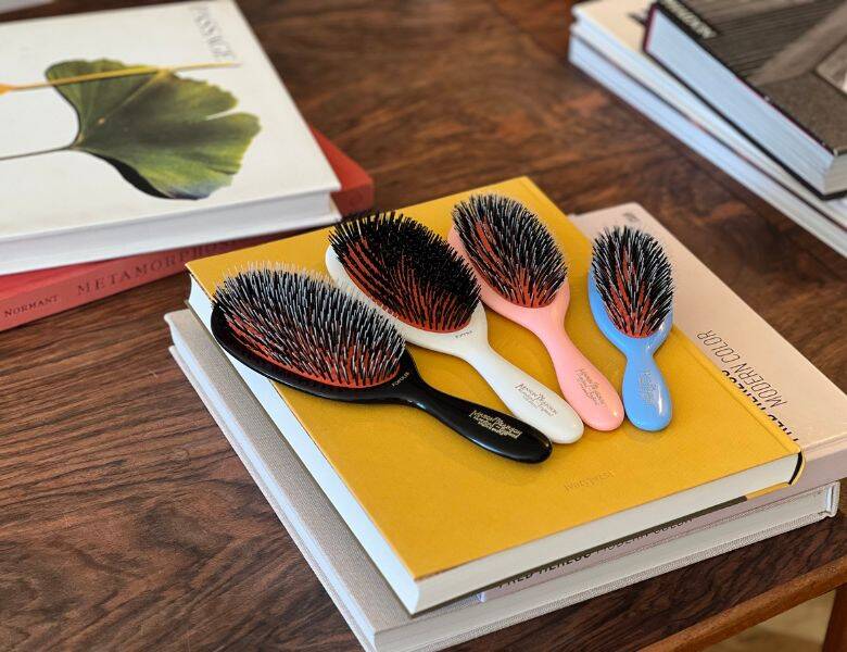 Her ses fire hårbørster i forskellige farver. De ligger på en gul bog, som ligger på et træbord med andre bøger omkring sig.