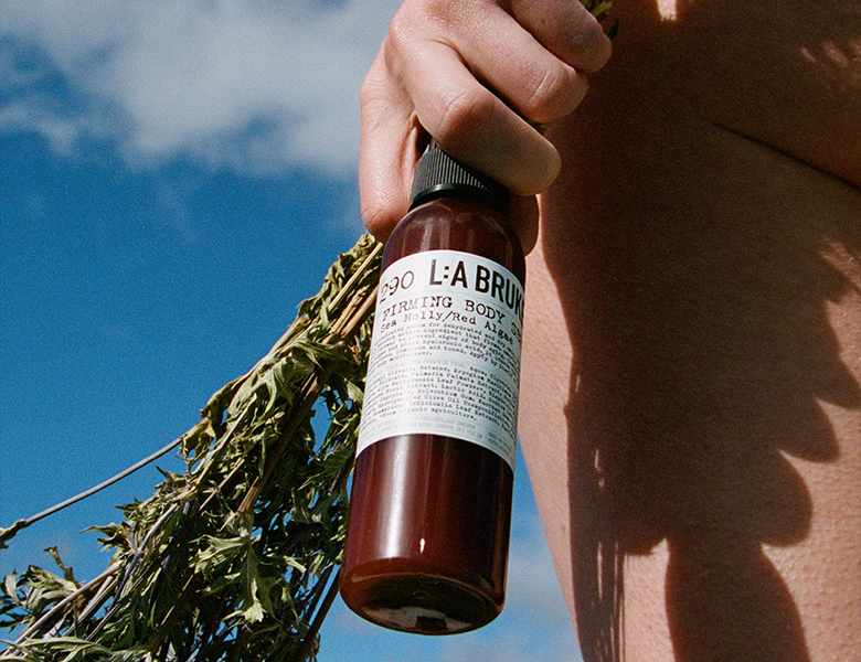 Her ses en hånd, der holder en L:A BRUKET flaske sammen med grene foran en blå himmel.