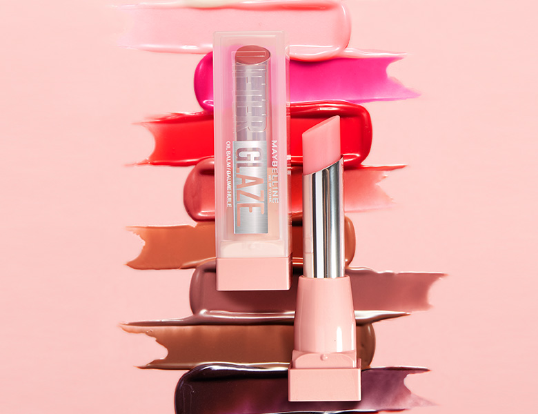 Her ses et billede af et læbeprodukt fra Maybelline placeret mod en lyserød baggrund med farvestrøg i forskellige nuancer. På produktet står teksten “MAYBELLINE LIFTER GLaze”. En åben lipstick er vist ved siden af emballagen.