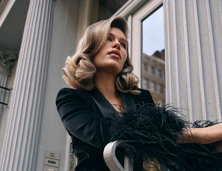 Her ses en kvinde med skulderlangt hår. Hun er beklædt i sort og står foran en grå bygning. 