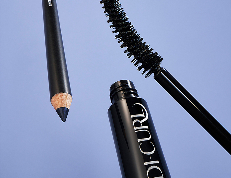 Her ses et billede med øjenmakeup-produkter placeret mod en blå baggrund. Der ses blandt andet en mascara med synlig børste og en eyelinerblyant.