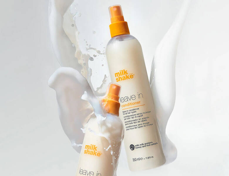 Her ses et billede med to flasker fra milk_shake placeret mod en lys baggrund med væske, der sprøjter omkring dem. På flaskerne står teksten “milk_shake leave in conditioner”. Flaskerne er i lyse nuancer med orange sprayhoveder.