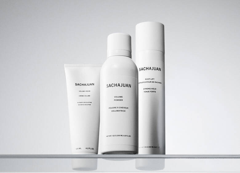 Her ses et billede med tre hårstylingprodukter fra SACHAJUAN placeret på en lys, reflekterende overflade mod en lys baggrund. På produkterne står teksten “SACHAJUAN”, “Volume Cream”, “Volume Powder” og “Root Lift”. Produkterne er i hvide emballager.