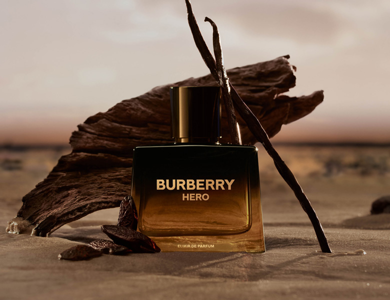 Her ses et billede med en parfumeflakon fra Burberry Goddess placeret foran en ravfarvet sten sammen med vaniljestænger og lilla blomster mod en varm beige baggrund. På flasken står blandt andet “Amber Vanilla”.
